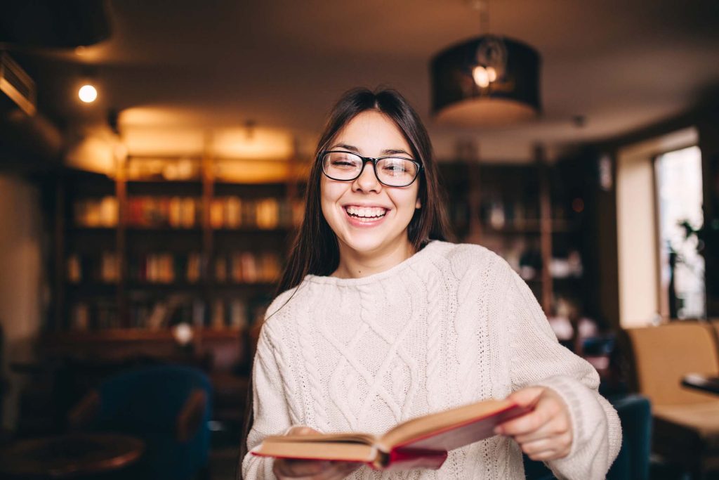 student-girl-laughing-and-holding-a-book-in-librar-EA5RJ2H.jpg